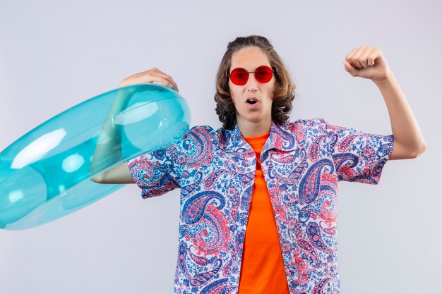 Young handsome guy wearing red sunglasses holding inflatable ring raising fist after a victory exited and happy standing 