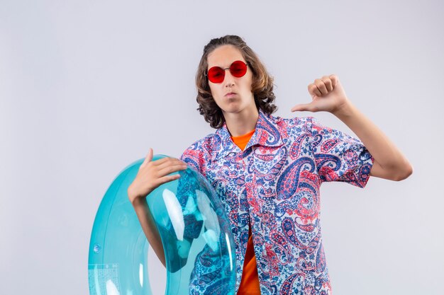 Young handsome guy wearing red sunglasses holding inflatable ring self-satisfied pointing to himself with thumb standing 