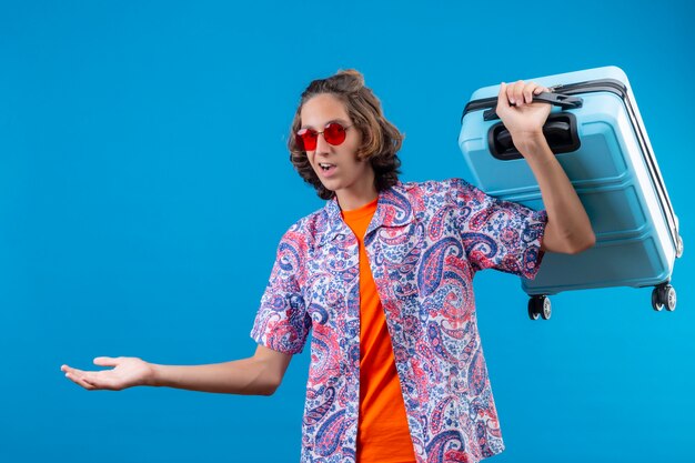 Young handsome guy wearing red sunglasses holding travel suitcase clueless and confused  having no answer spreading arms standing over blue background