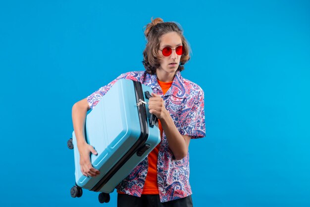 青い背景の上に立って混乱している旅行スーツケースを持って赤いサングラスをかけている若いハンサムな男