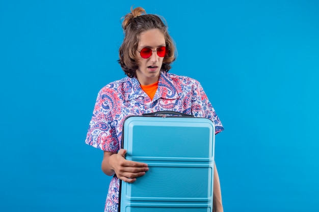 Бесплатное фото Молодой красивый парень в красных солнцезащитных очках держит чемодан для путешествий и выглядит смущенным стоя