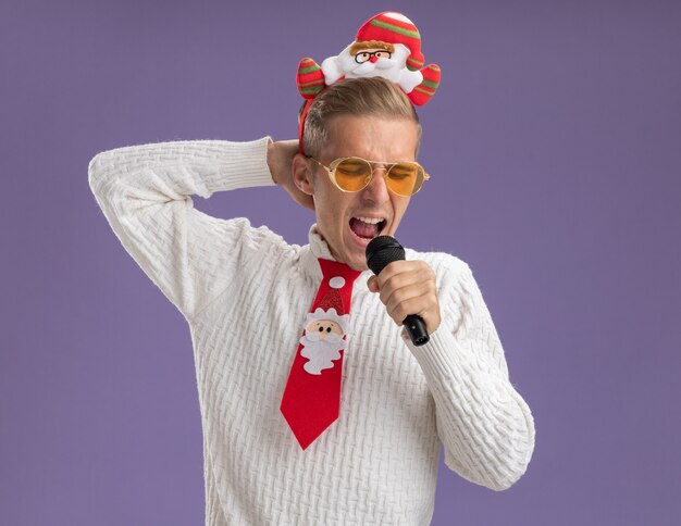 Young handsome guy wearing santa claus headband and tie with glasses holding microphone keeping hand behind head singing with closed eyes isolated on purple background