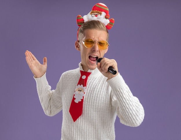 Young handsome guy wearing santa claus headband and tie with glasses holding microphone showing empty hand singing with closed eyes isolated on purple background