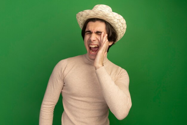 Young handsome guy with closed eyes wearing hat calling someone isolated on green wall