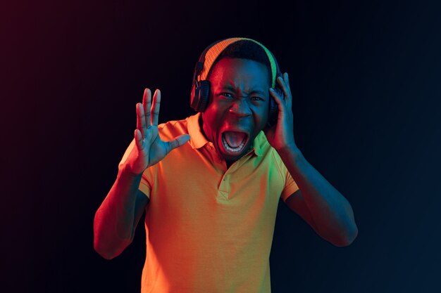 The young handsome happy hipster man listening music with headphones at black studio with neon lights. Disco, night club, hip hop style, positive emotions, face expression, dancing concept