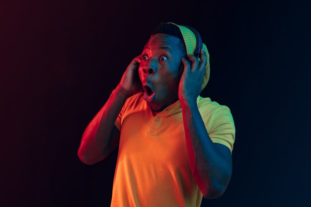 The young handsome happy hipster man listening music with headphones at black studio with neon lights. Disco, night club, hip hop style, positive emotions, face expression, dancing concept