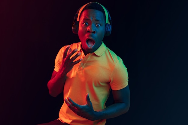 The young handsome happy hipster man listening music with headphones at black studio with neon lights. Disco, night club, hip hop style, positive emotions, face expression, dancing concept