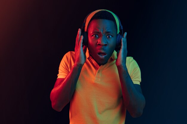 The young handsome happy hipster man listening music with headphones at black studio with neon lights. Disco, night club, hip hop style, positive emotions, face expression, dancing concept