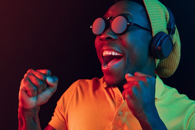 The young handsome happy hipster man listening music with headphones at black studio with neon lights. Disco, night club, hip hop style, positive emotions, face expression, dancing concept