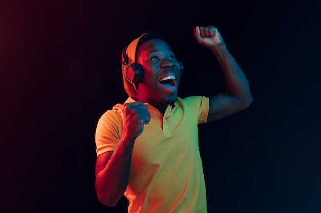 The young handsome happy hipster man listening music with headphones at black studio with neon lights. Disco, night club, hip hop style, positive emotions, face expression, dancing concept