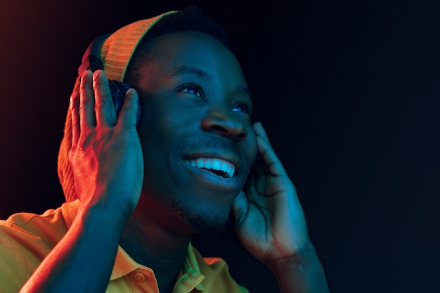 The young handsome happy hipster man listening music with headphones at black studio with neon lights. Disco, night club, hip hop style, positive emotions, face expression, dancing concept