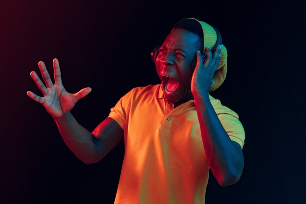 The young handsome happy hipster man listening music with headphones at black studio with neon lights. Disco, night club, hip hop style, positive emotions, face expression, dancing concept