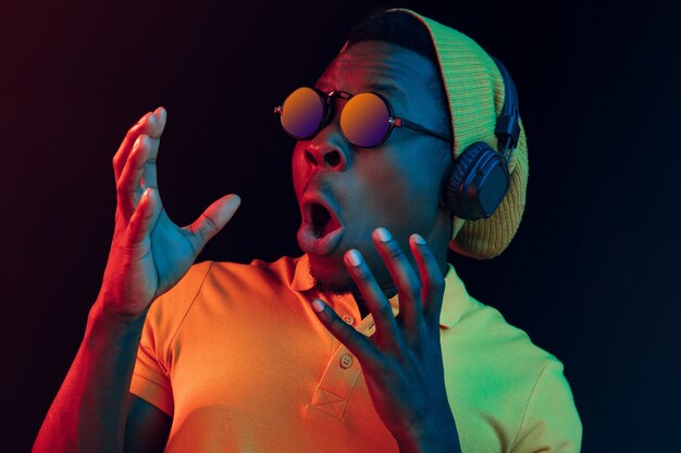 The young handsome happy hipster man listening music with headphones at black studio with neon lights. Disco, night club, hip hop style, positive emotions, face expression, dancing concept