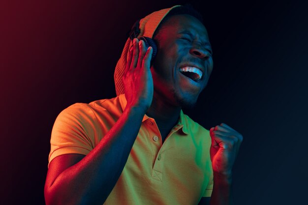 The young handsome happy hipster man listening to music with headphones at black studio with neon lights