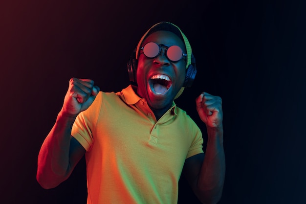 The young handsome happy hipster man listening to music with headphones at black studio with neon lights