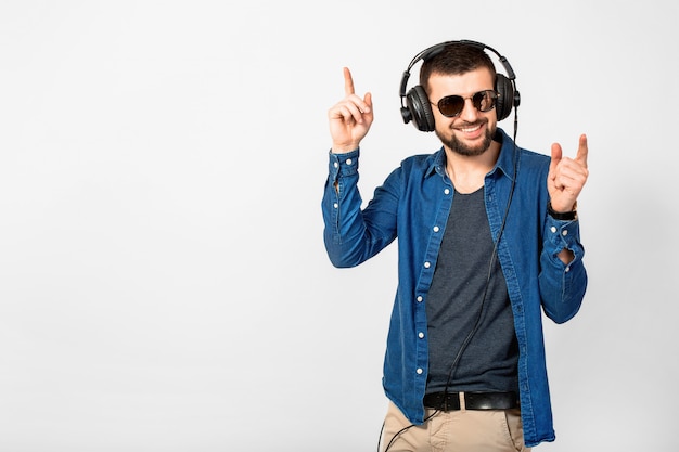 Молодой красивый счастливый улыбающийся человек танцует и слушает музыку в наушниках на белом фоне студии, в джинсовой рубашке и солнцезащитных очках