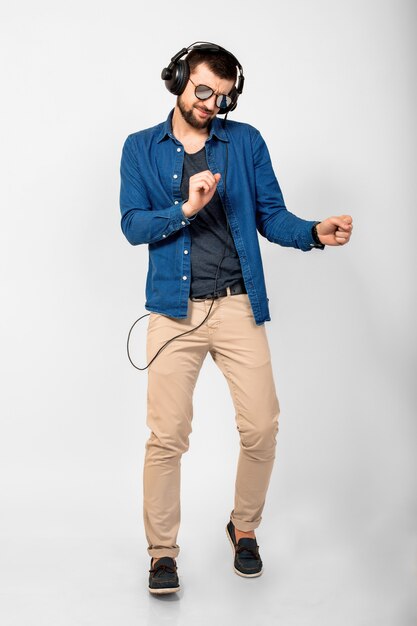 Молодой красивый счастливый улыбающийся человек танцует и слушает музыку в наушниках на белом фоне студии, в джинсовой рубашке и солнцезащитных очках