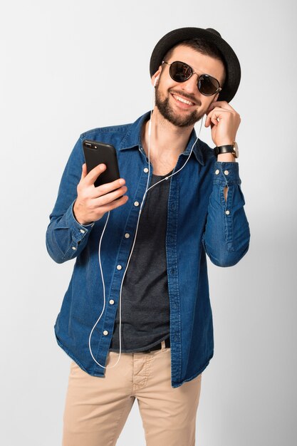 Young handsome happy smiling man listening to music in earphones isolated on white studio background, holding smartphone, wearing denim shirt, hat and sunglasses