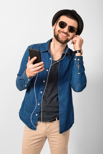 Молодой красивый счастливый улыбающийся человек, слушающий музыку в наушниках, изолированные на белом фоне студии, держа смартфон, одетый в джинсовую рубашку, шляпу и солнцезащитные очки