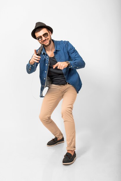 Young handsome happy smiling man listening to music in earphones isolated on white studio background, holding smartphone, wearing denim shirt, hat and sunglasses