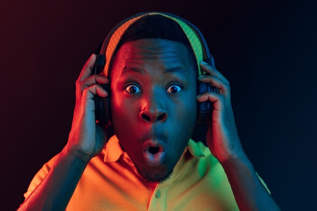 The young handsome happy surprised hipster man listening music with headphones at black studio with neon lights. Disco, night club, hip hop style, positive emotions, face expression, dancing concept