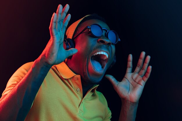 The young handsome happy surprised hipster man listening music with headphones at black studio with neon lights. Disco, night club, hip hop style, positive emotions, face expression, dancing concept