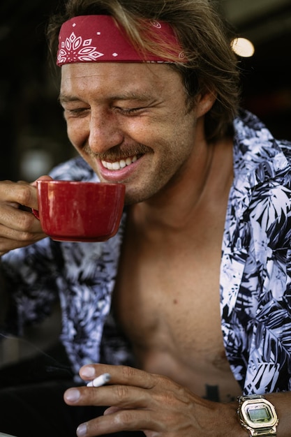 Молодой красивый мужчина-хиппи в красной бандане с обнаженным торсом в кафе на Бали. Счастливый человек улыбается