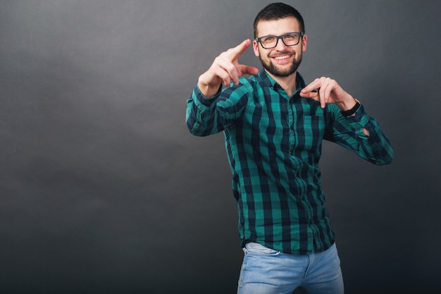 Young handsome hipster bearded man on grey background, green checkered shirt, eyeglasses, pointing finger, happy, smiling, positive emotion