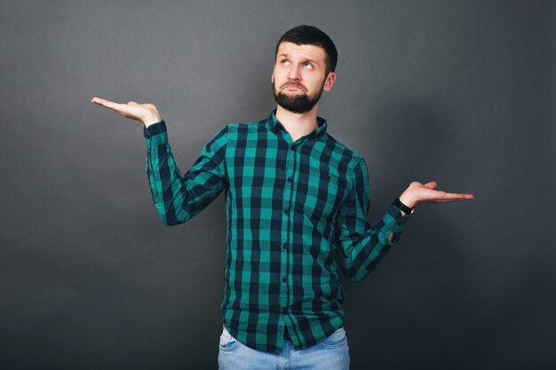 Young handsome hipster bearded man holding hands up evaluation question, green checkered shirt, grey background