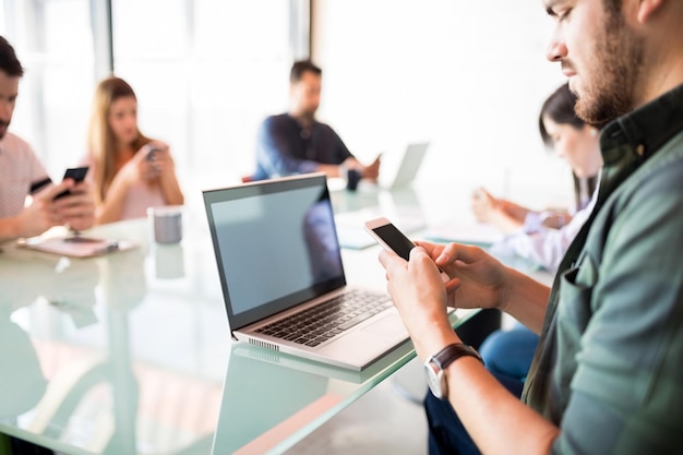 Free photo young handsome latin man sitting at conference table using mobile phone with colleagues sitting by