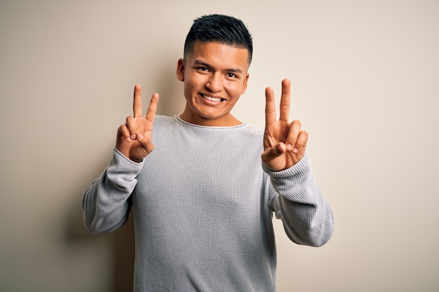 Foto gratuita giovane bell'uomo latino che indossa un maglione casual in piedi su sfondo bianco isolato sorridente guardando la telecamera mostrando le dita facendo segno di vittoria numero due