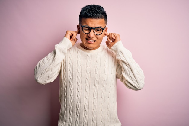 Young handsome latin man wearing white casual sweater and glasses over pink background covering ears with fingers with annoyed expression for the noise of loud music Deaf concept