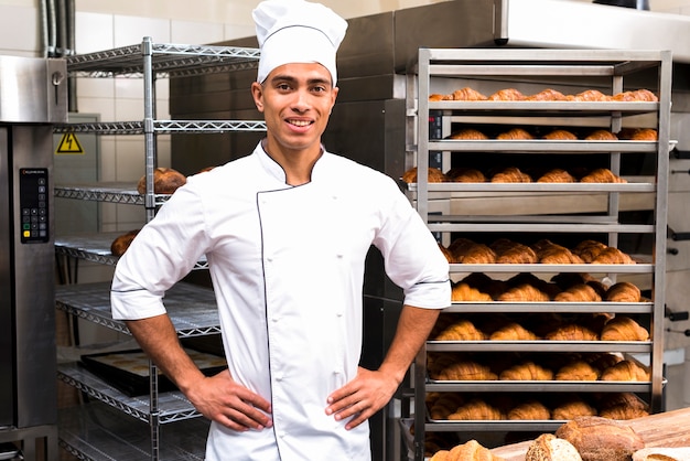 Young handsome male baker in white uniform standing against baking shelf