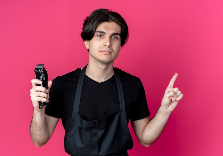 मुफ़्त फोटो young handsome male barber in uniform holding hair clippers and points at up isolated on pink wall
