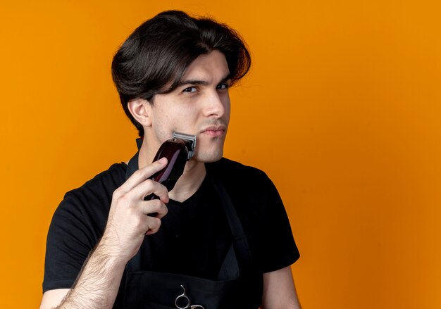 young handsome male barber in uniform trimming his beard with hair clippers isolated on orange wall