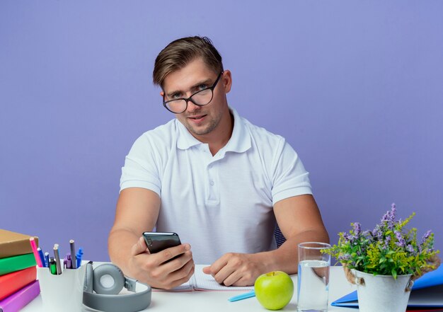 young handsome male student sitting at desk with school tools wearing glasses and holding phone isolated on blue wall