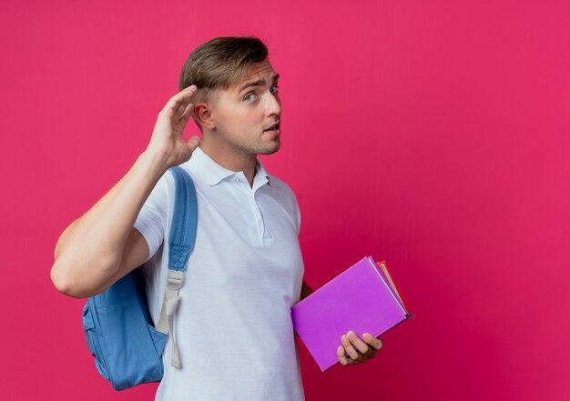 young handsome male student wearing backpack holding books and showing listen gesture 