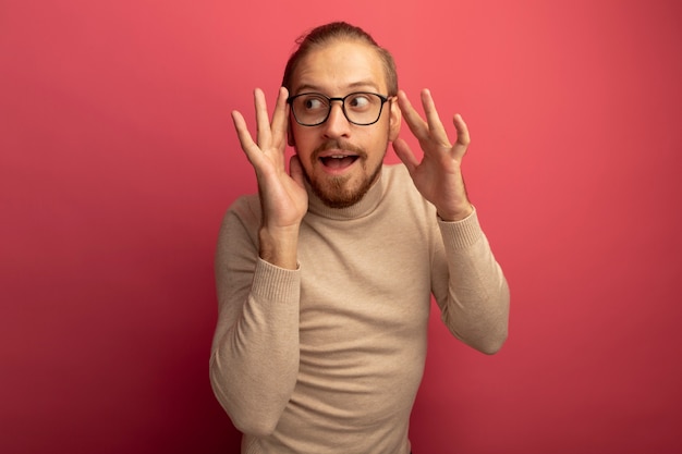 Young handsome man in beige turtleneck and glasses looking aside smiling slyly standing over pink wall