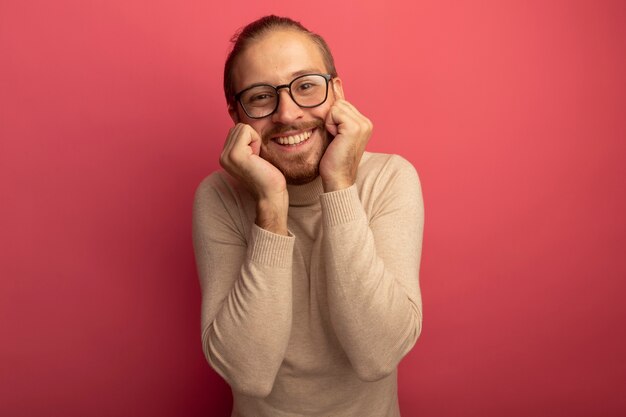 베이지 색 터틀넥과 안경에 젊은 잘 생긴 남자가 분홍색 벽 위에 서있는 긍정적 인 감정을 유쾌하게 느끼고 웃고 있습니다.