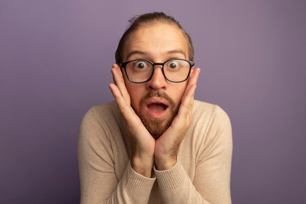 Young handsome man in beige turtleneck and glasses surprised and amazed 