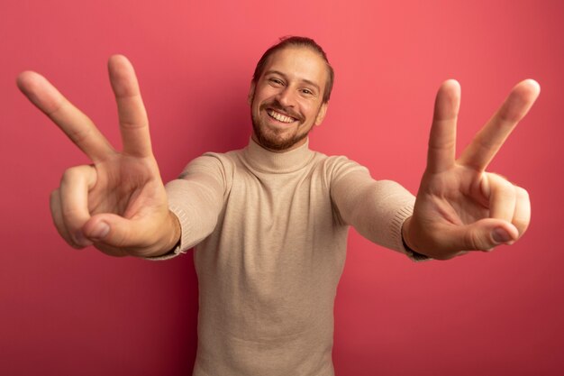 Young handsome man in beige turtleneck looking at front smiling cheerfully showing v-sign with both hands standing over pink wall