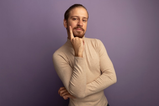 Young handsome man in beige turtleneck looking at front with hand on his chin thinking standing over lilac wall