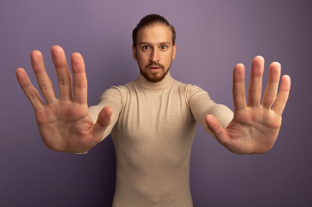 Young handsome man in beige turtleneck looking at front worried making stop gesture with open hands standing over lilac wall
