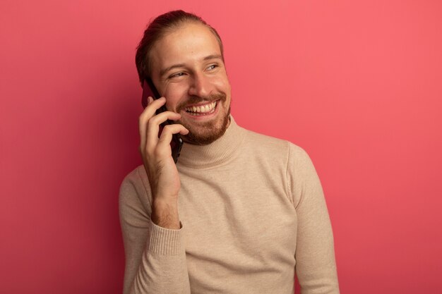 ピンクの壁の上に立っている携帯電話で話しながら元気に笑っているベージュのタートルネックの若いハンサムな男