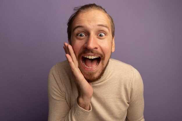 Young handsome man in beige turtleneck surprised with hand near mouth 