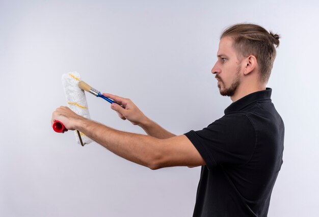 Young handsome man in black polo shirt holding paint roller painting it with a paint brush with serious face standing over white background