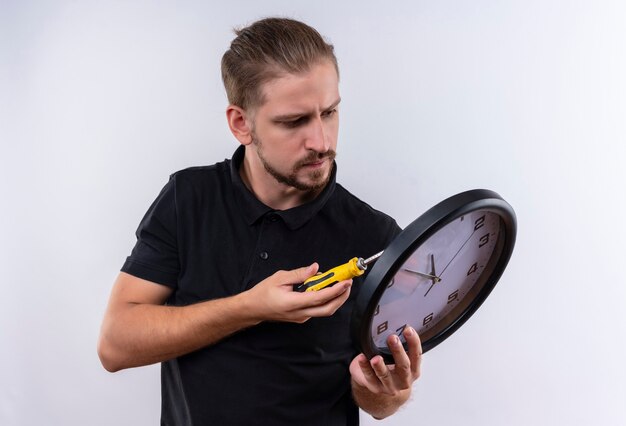 Free photo young handsome man in black polo shirt holding wall clock reparing it with serious face standing over white background