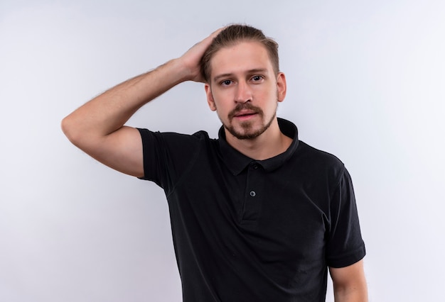 Young handsome man in black polo shirt looking confused touching head standing over white background