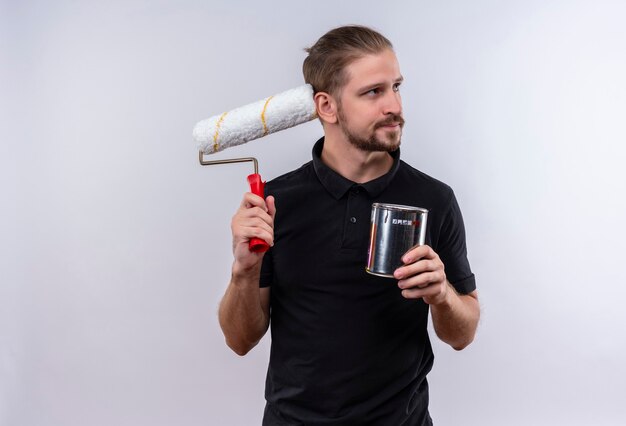 Young handsome man in black polo shirt scratching head with a paint roller holding paint can looking aside with skeptic expression standing over white background