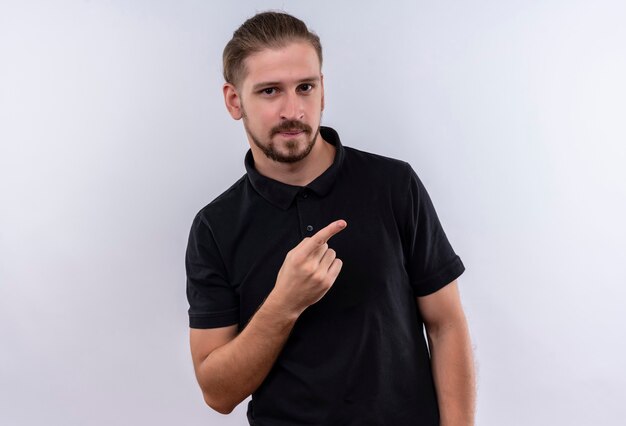 Young handsome man in black polo shirt smiling confident pointing to the side with finger standing over white background